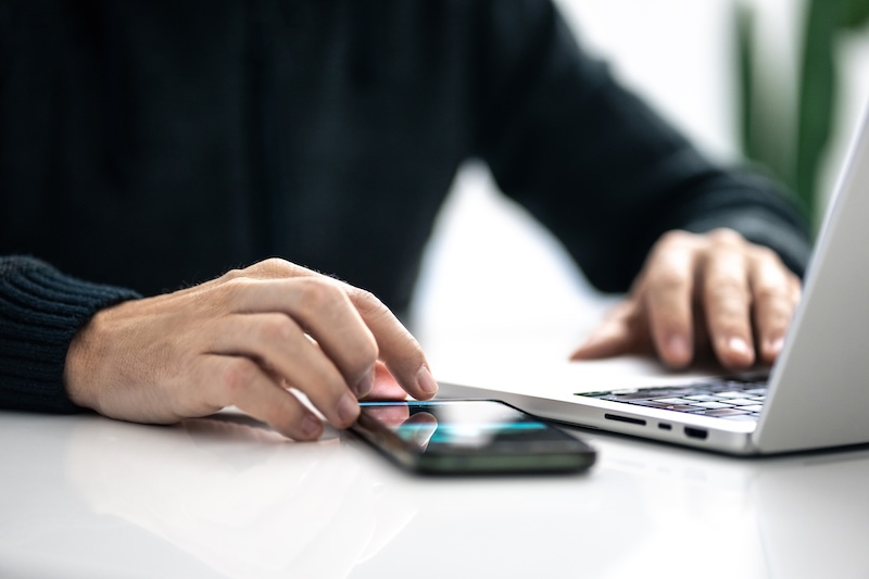 A person wearing a dark sweater uses a laptop with one hand while touching a smartphone on a white desk with the other, exploring internet security tools.