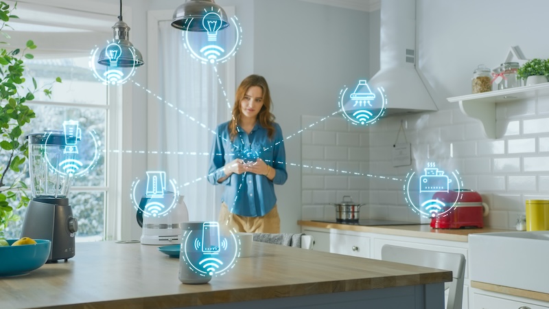 A woman stands in a modern kitchen using a smartphone, with illustrated icons showing smart home appliances connected wirelessly—highlighting the convenience and internet of things privacy concerns of linked lights, fridge, thermostat, speaker, and more.