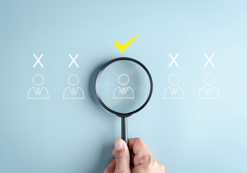 A hand holds a magnifying glass over one of four outlined human icons, symbolizing selection—like learning how to remove items from LexisNexis—with a yellow check mark above the chosen icon, while the others display white X marks.
