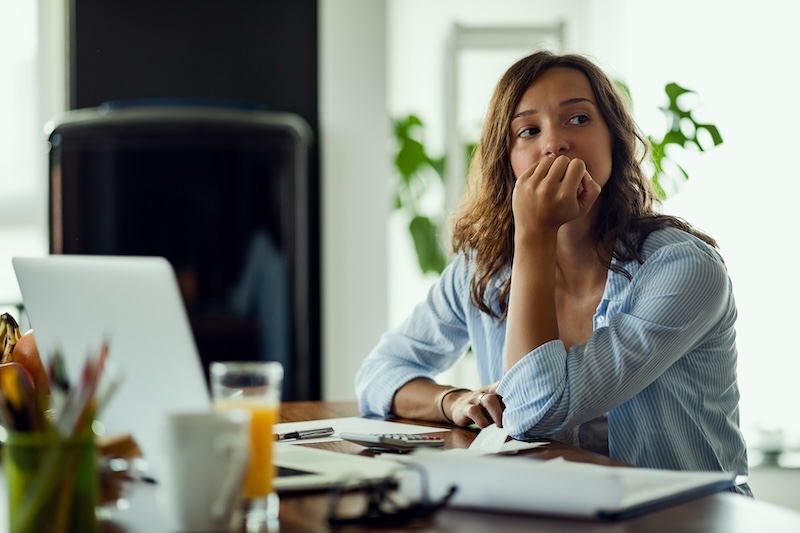 A woman sits at a desk with a laptop and papers, resting her chin on her hand and thinking about how to remove items from LexisNexis. The workspace is bright, with plants and a glass of orange juice nearby.