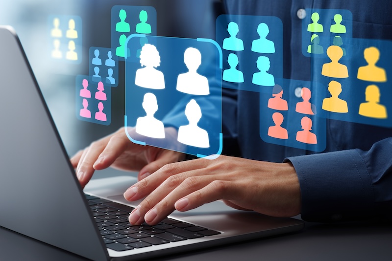 Hands typing on a laptop keyboard with digital icons of people and groups floating above, representing social networks, online communication, virtual teams, or efforts to block TruthFinder.