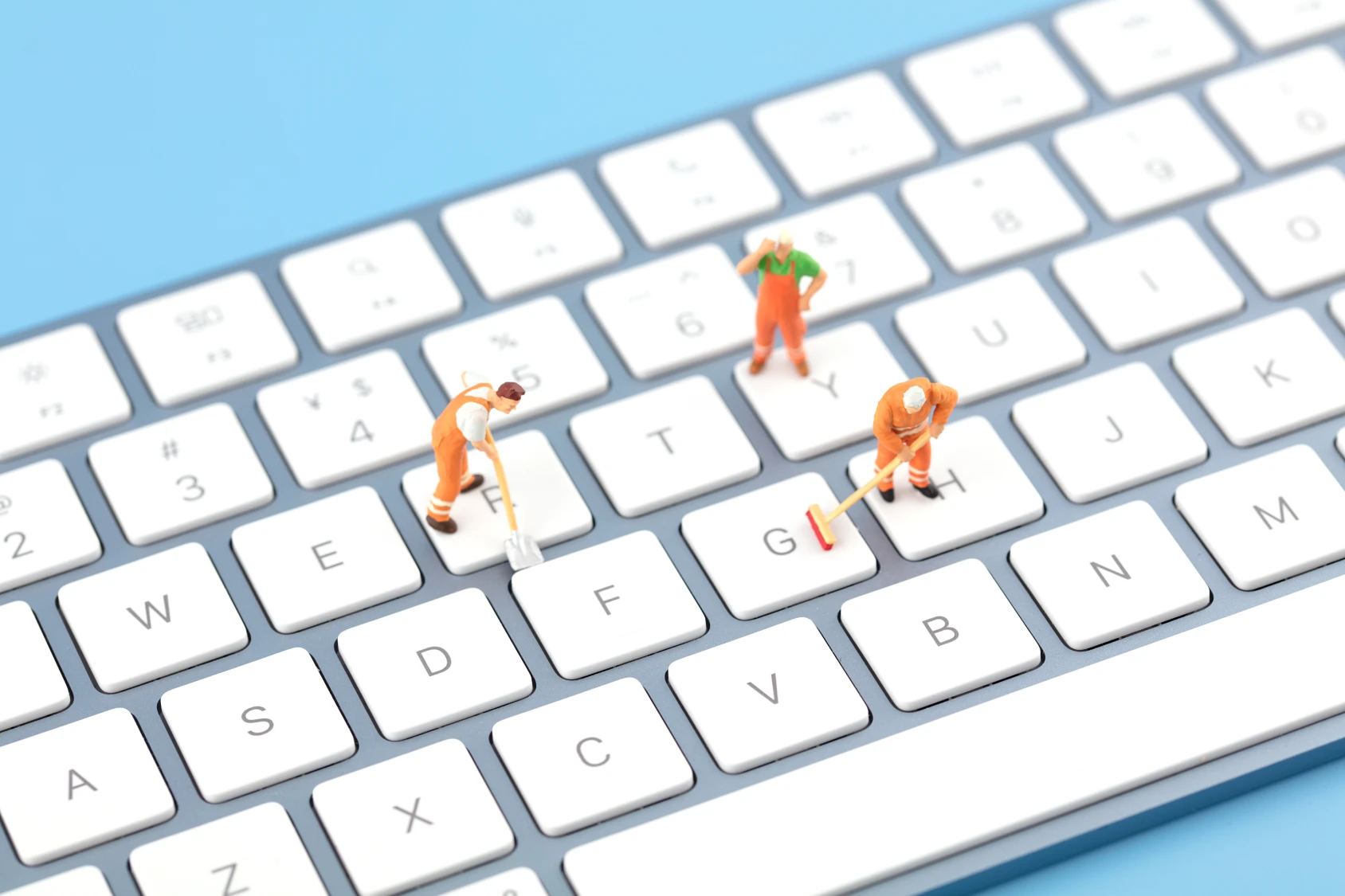 Miniature figures dressed as construction workers are on a white computer keyboard. One figure appears to be sweeping, another is standing and observing, while the third is using a tool, symbolizing reputation cleanup. The background is a light blue surface.