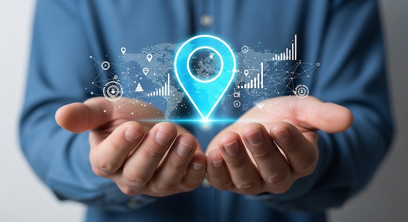 A person in a blue shirt holds out cupped hands displaying a glowing digital location pin surrounded by data graphs, network lines, and a world map, symbolizing global connectivity and advanced location tracking technology.