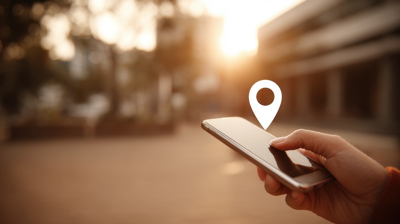 A hand holds a smartphone outdoors at sunset, with a digital map pin icon hovering above the screen, symbolizing GPS or location tracking services.