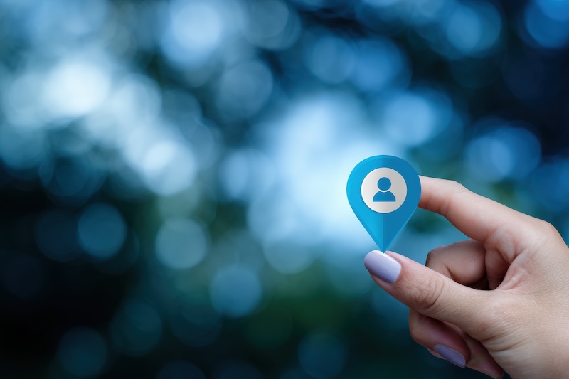 A hand with light purple nail polish holds a blue map pin icon featuring a white user symbol, representing Location Tracking, against a blurred outdoor background with blue and white bokeh lights.