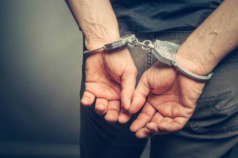 A person in a gray shirt and pants is shown from behind with their hands handcuffed behind their back, evoking the look of an arrest, often seen when searching how to find someone's mugshots. The background is neutral and out of focus.