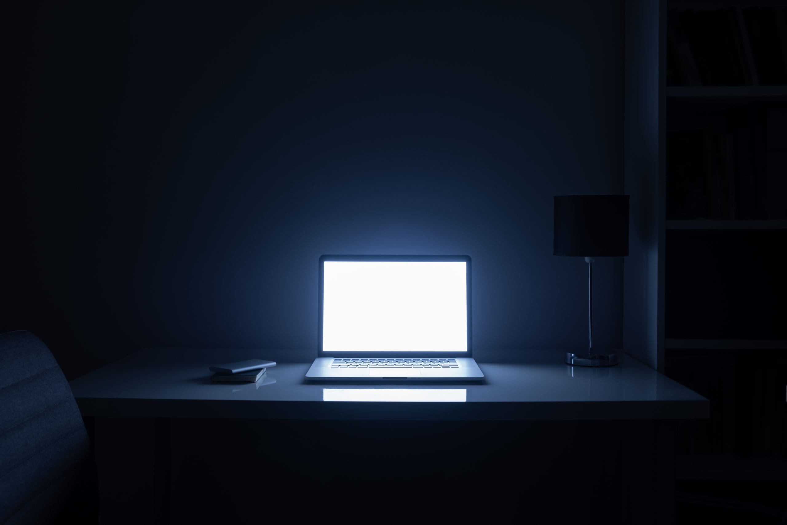 A laptop open with a blank white screen illuminating a dimly lit room, casting a blue glow over a minimalist workspace at night.