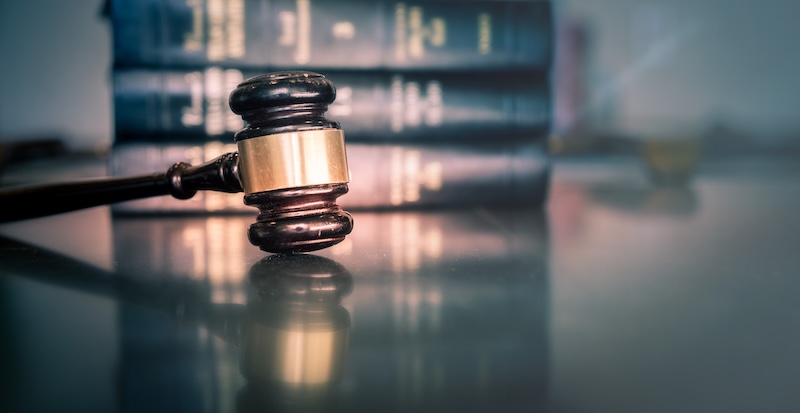 A judge’s gavel rests on a reflective surface with blurred legal books and voterrecords stacked in the background, symbolizing law and justice.