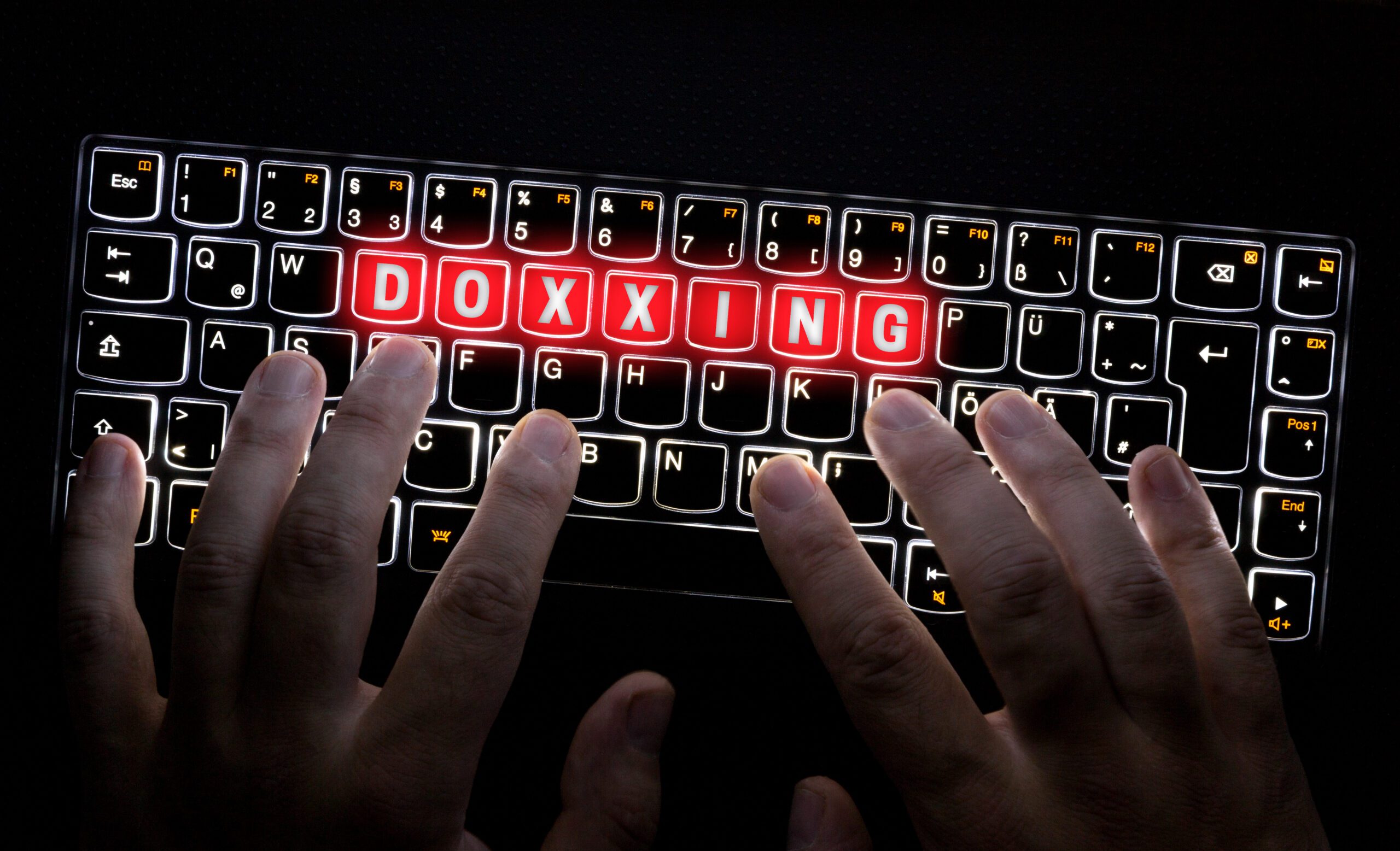 Hands typing on a backlit keyboard with the word "doxxing" illuminated in red letters on the keys.