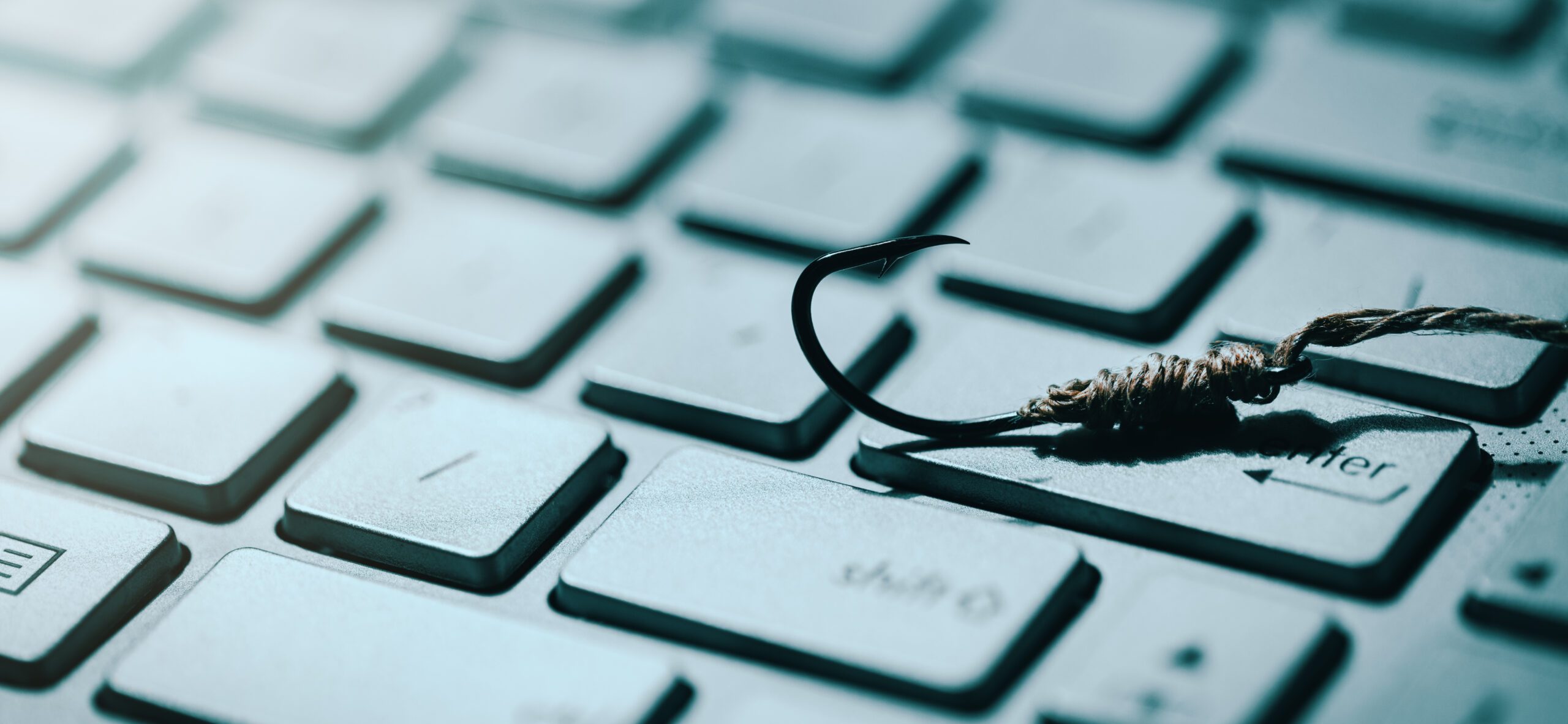 A fishing hook lies on a computer keyboard, symbolizing the concept of phishing in cybersecurity. The hook is prominently placed on the "Enter" key amidst the white keys, highlighting the threat of online scams and deceptive tactics.