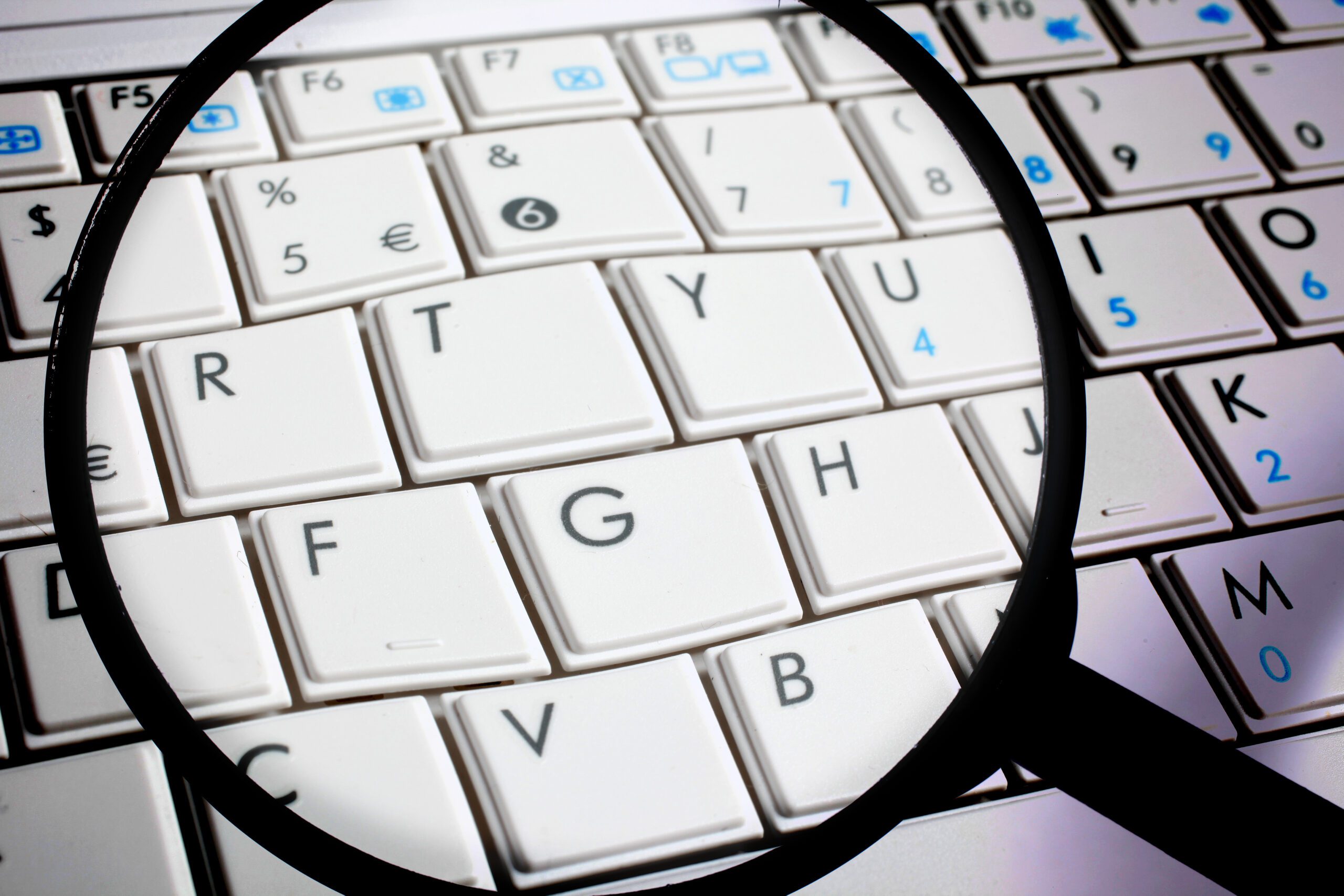 A close-up image of a white computer keyboard with a black magnifying glass placed over it. The magnifying glass highlights the keys "T," "Y," "G," "H," "V," and "B," making them appear larger. The keys have various symbols and letters on them.