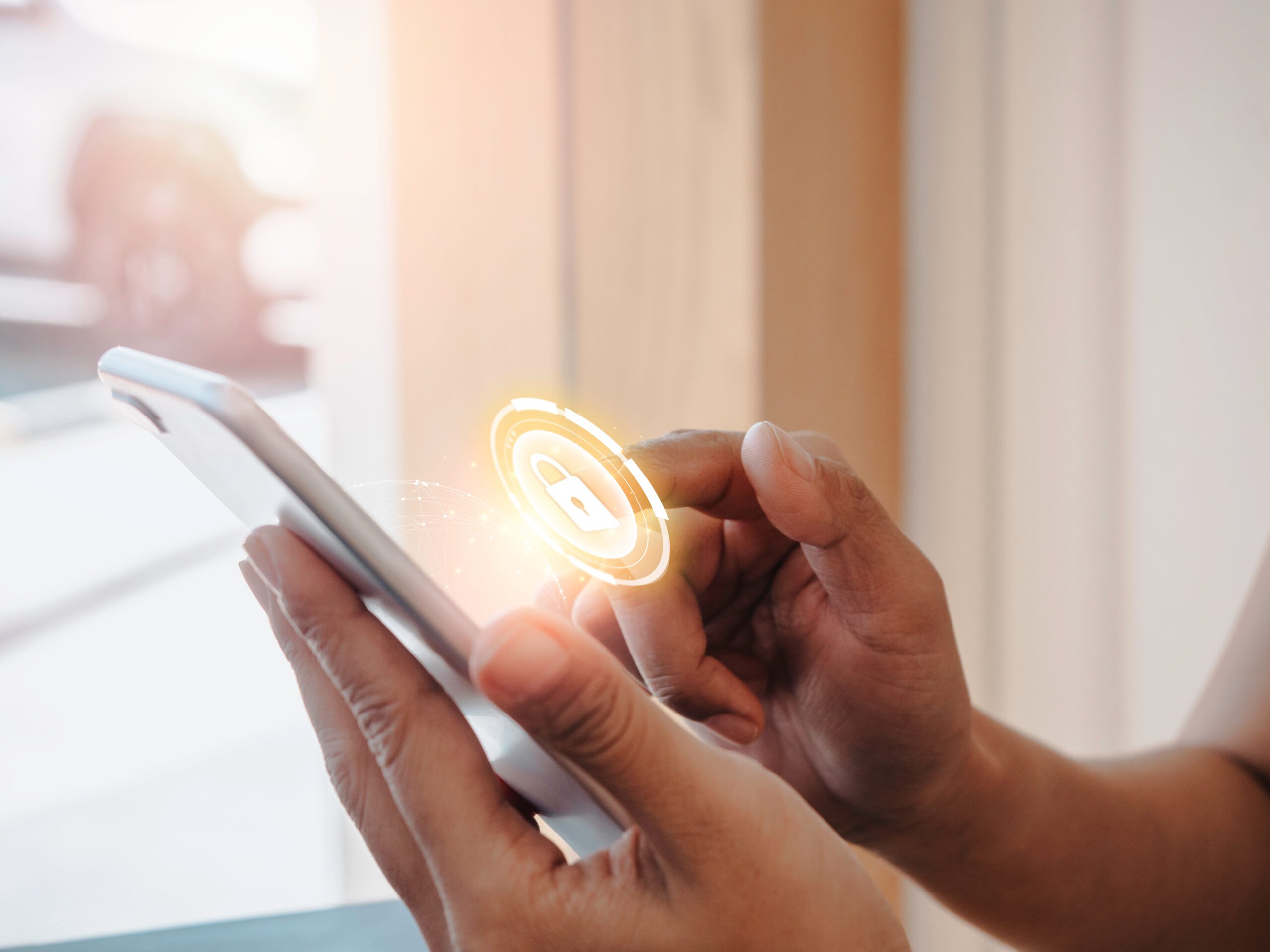 A person's hands holding a smartphone with an illuminated lock icon hovering above it, signifying security or authentication. The bright light from the window suggests a daytime setting.