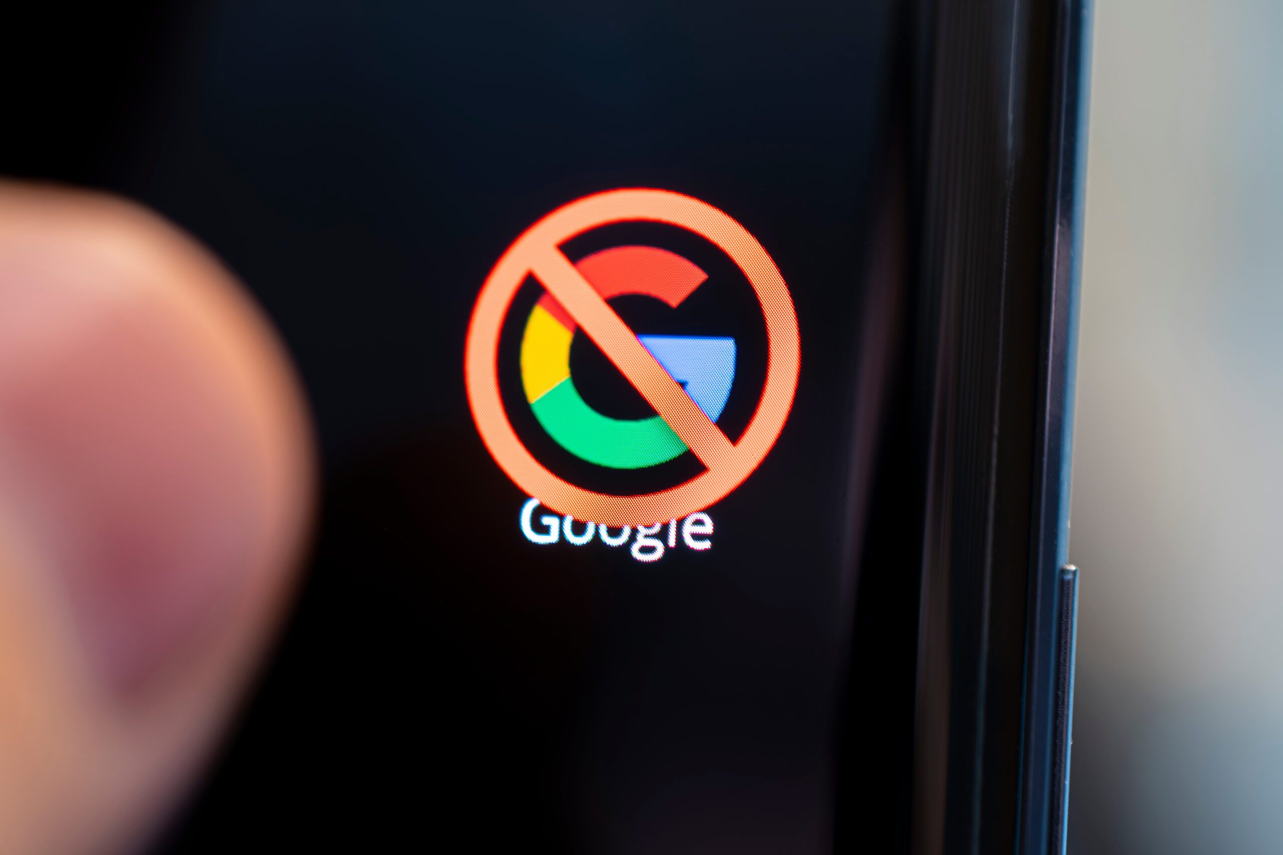 A smartphone screen displaying the Google logo with a red prohibition symbol over it, indicating a blocked or restricted access to the service. The text "Google" is visible below the logo. A finger is partially visible on the left side.