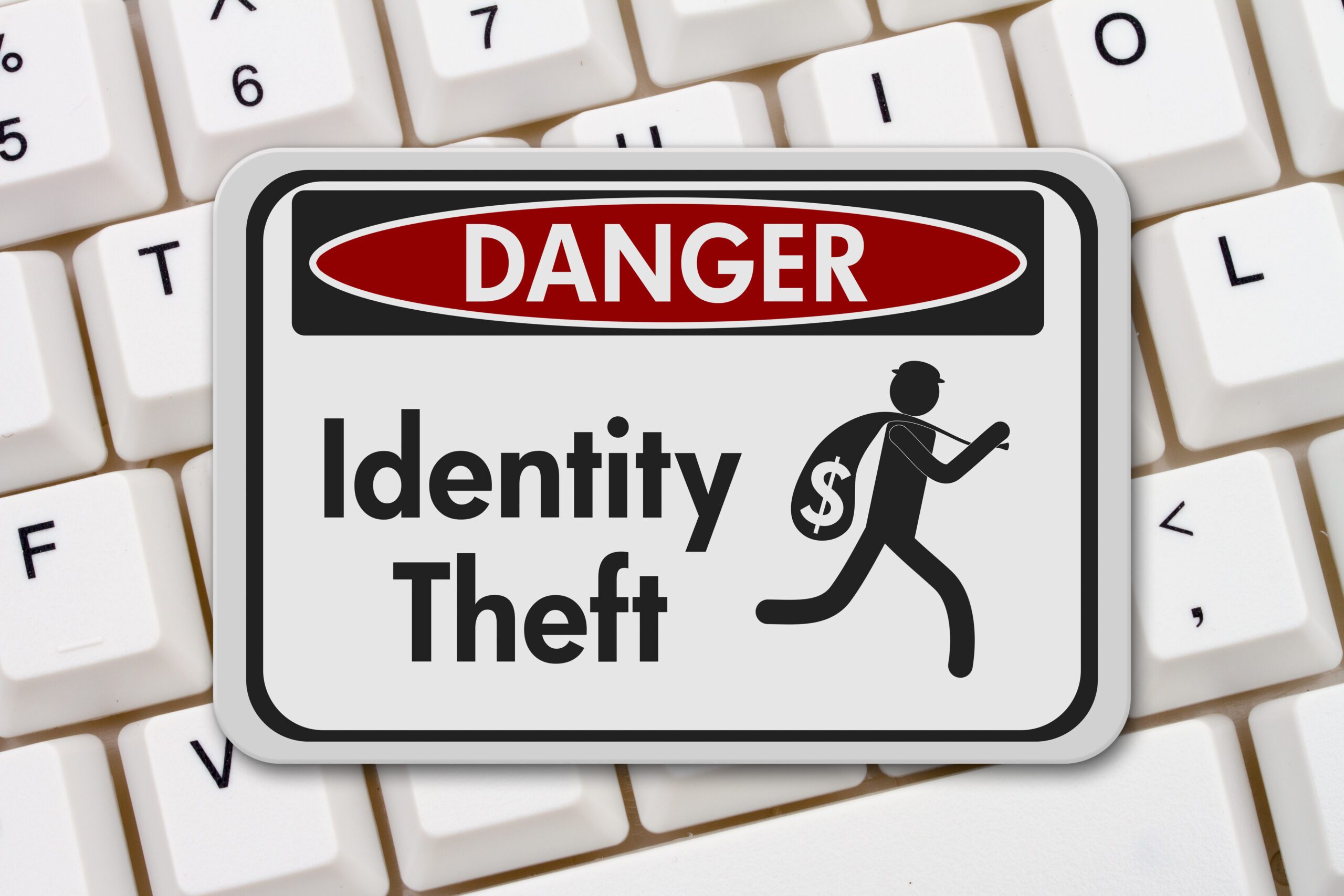 A warning sign titled "Danger Identity Theft" is displayed against a computer keyboard background. The sign includes an icon of a person running with a sack marked with a dollar symbol, symbolizing theft.