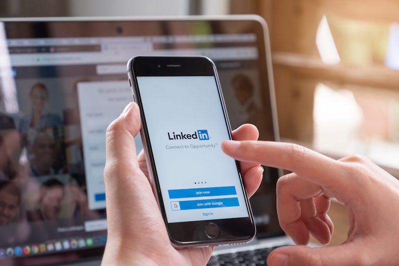 A person holds a smartphone displaying the LinkedIn login screen, with a laptop showing blurred profiles in the background—highlighting concerns about LinkedIn identity theft as the user touches the screen to sign in.