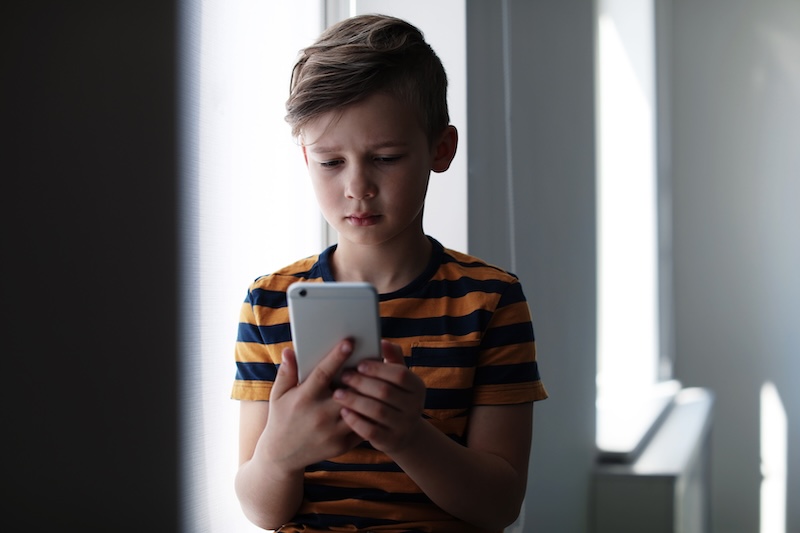 A young boy wearing a striped t-shirt sits by a window, looking intently at a smartphone he is holding with both hands. Sunlight streams in, softly illuminating his serious expression as he explores internet safety for kids.