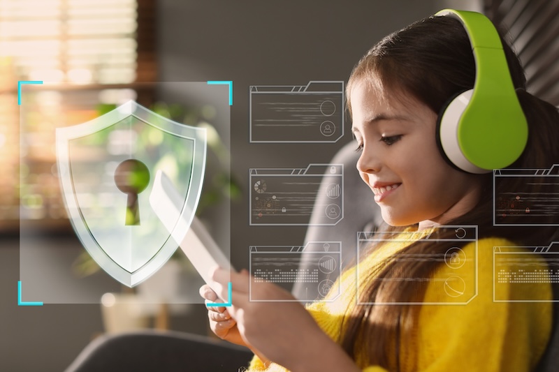 A smiling girl wearing green headphones uses a tablet. A digital shield with a keyhole and virtual icons are superimposed, highlighting internet safety for kids and the importance of cybersecurity and online protection for children.