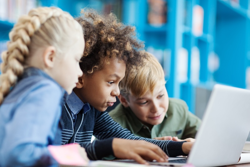 Three children sit closely together, focused on a laptop screen in a brightly lit room with blue shelves in the background, learning about internet safety for kids.
