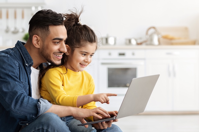 A man and a young girl sit together in a bright kitchen, smiling at a laptop screen. The girl points as the man types, showing they’re enjoying something online while practicing internet safety for kids.