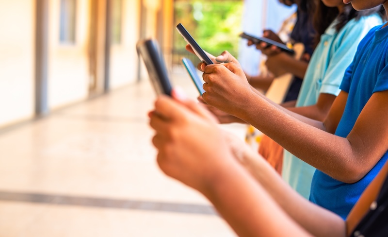 A row of young people holding and using smartphones, standing indoors with their faces out of frame, focused on their devices—highlighting the importance of internet safety for kids in today’s connected world.