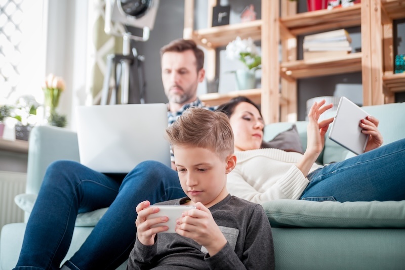 A boy sits on a sofa using a smartphone, practicing internet safety for kids, while a woman reclines next to him with a tablet and a man in the background works on a laptop. All are focused on their devices in a cozy living room.