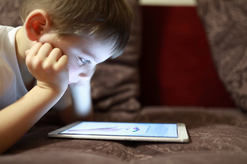 A young boy lies on a couch, intently looking at a tablet screen in front of him. He rests his chin on his hands, the softly lit room highlighting his focused expression as he explores internet safety for kids.