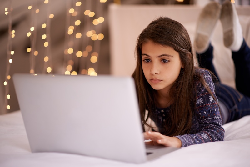 A young girl with long brown hair lies on her stomach on a bed, using a laptop. She looks focused, practicing internet safety for kids. Warm fairy lights are blurred in the background, creating a cozy atmosphere.
