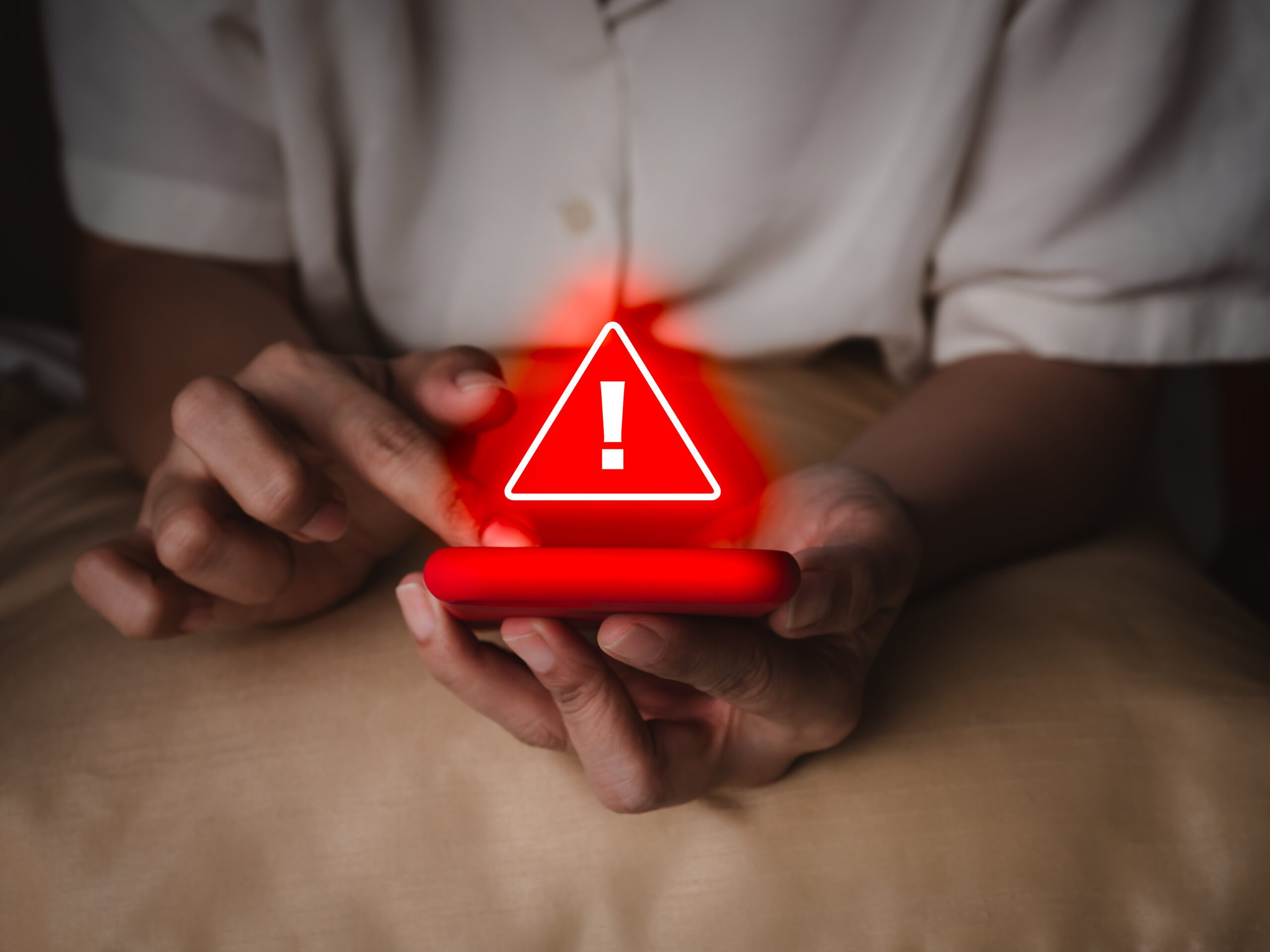 A person holding a smartphone with a glowing red warning icon and exclamation mark appearing above the screen, suggesting an alert or security warning.