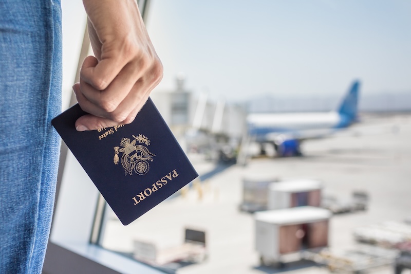A person holds a U.S. passport in their hand while standing by an airport window, with a parked airplane and airport equipment visible outside on the tarmac.